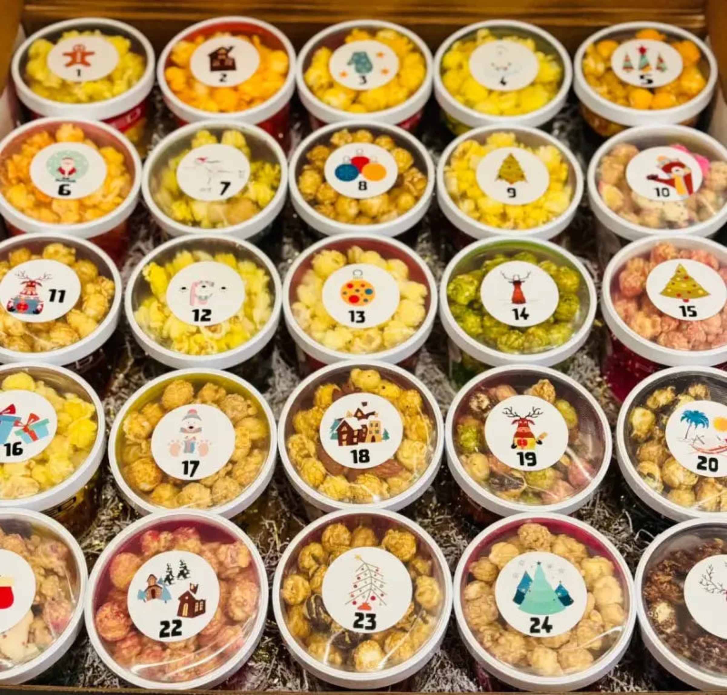 An advent calendar box filled with twenty-four individual tubs of gourmet popcorn, each with a unique Christmas-themed sticker.
