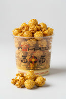 A clear plastic cup filled with caramel popcorn and topped with extra popcorn pieces, with a few popcorn kernels placed in front on a light gray background. The cup has 'POPPED 505' branding on the front.