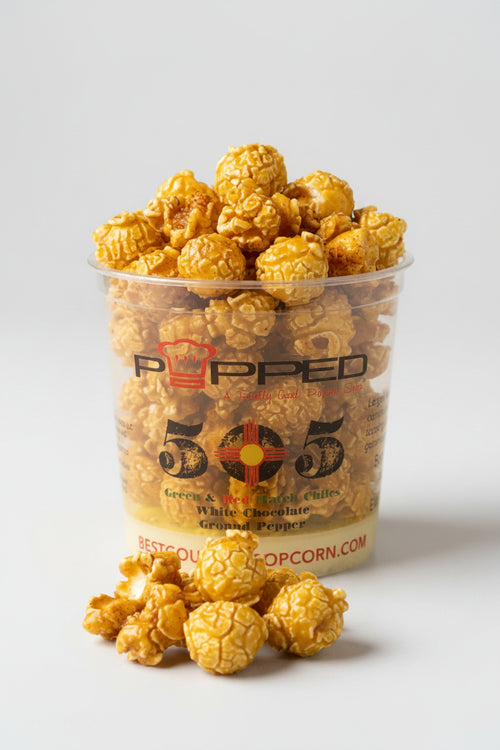 A clear plastic cup filled with caramel popcorn and topped with extra popcorn pieces, with a few popcorn kernels placed in front on a light gray background. The cup has 'POPPED 505' branding on the front.