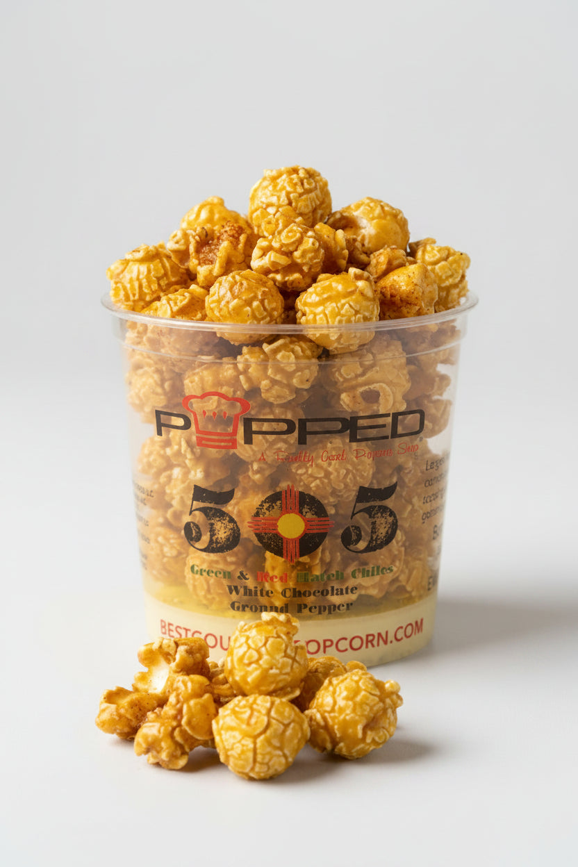 A clear plastic cup filled with caramel popcorn and topped with extra popcorn pieces, with a few popcorn kernels placed in front on a light gray background. The cup has 'POPPED 505' branding on the front.