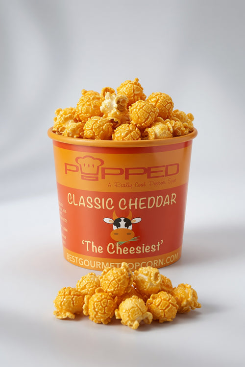 A container of Popped Classic Cheddar popcorn with several pieces spilled in front, set against a clean white studio background. The container features a bright orange label with a cow illustration and the brand name 'Popped'.