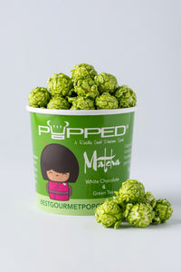 A green cup of Popped Matcha green chocolate popcorn with several popcorn balls placed in front on a light gray background.