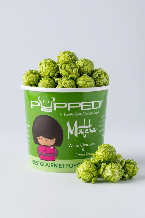 A green cup of Popped Matcha green chocolate popcorn with several popcorn balls placed in front on a light gray background.