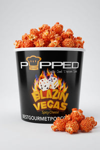 A black bucket of Popped Blazin Vegas Spicy Cheese popcorn, filled with orange-red popcorn and with a few pieces scattered in front, on a light gray background.