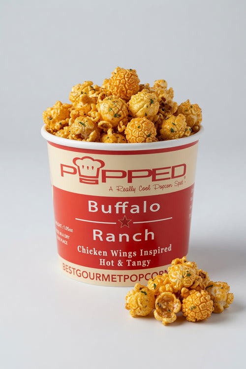 A red and cream paper cup of Popped Buffalo Ranch popcorn filled with golden, crispy popcorn kernels, with a few kernels scattered in front on a light gray background.