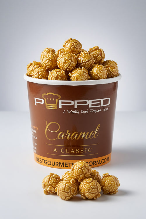 A brown paper cup filled with caramel popcorn, with several popcorn kernels placed in front on a light gray studio background. The cup is branded Popped with the text 'Caramel A Classic' and 'Best Gourmet Popcorn'.