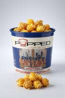 A container of Popped Chi City Mix Sauté & Cheese popcorn, filled with golden-brown popcorn and with a small pile in front, on a light gray background.