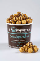 A cup of Popped Chocolate Tide popcorn filled with caramel and dark chocolate drizzled popcorn, with a few pieces spilled in front, on a light gray background.