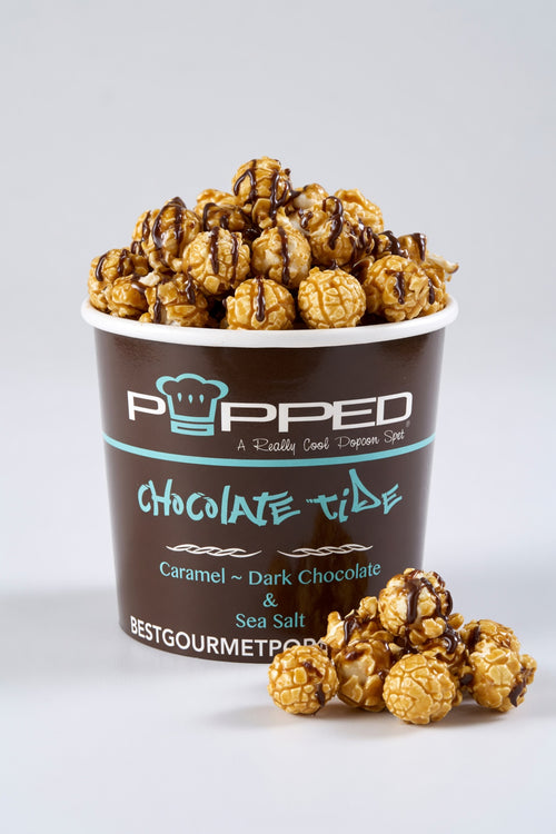 A cup of Popped Chocolate Tide popcorn filled with caramel and dark chocolate drizzled popcorn, with a few pieces spilled in front, on a light gray background.