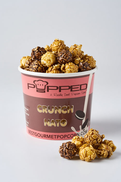 A cup of gourmet popcorn from Popped Creamery, featuring Crunch Mato flavor. The cup is filled with a mix of light caramel popcorn and dark chocolate-coated popcorn, with a few pieces spilled in front on a light gray background.