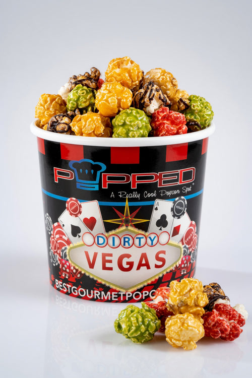 A cup of Popped Dirty Vegas gourmet popcorn filled with multicolored popcorn pieces, with a few pieces scattered in front on a light gray background.