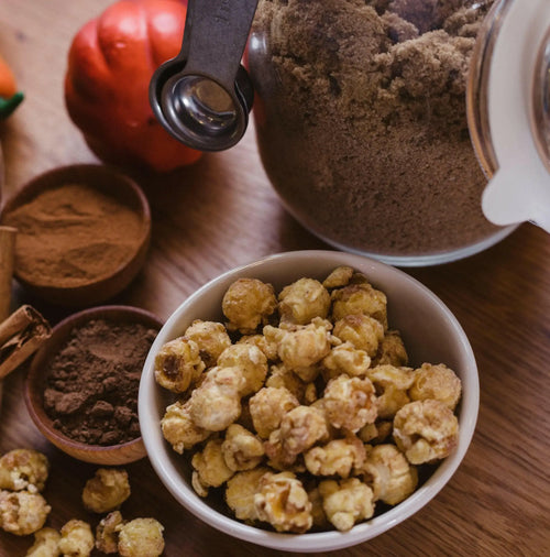 A bowl of caramel popcorn, surrounded by cinnamon sticks, cocoa powder, and a jar of brown sugar.