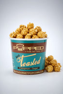 A cup of Popped Toasted Cinnamon + Sugar gourmet popcorn with caramel-coated popcorn piled over the rim and a few pieces resting beside it on a light gray studio background.