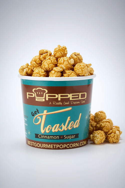 A cup of Popped Toasted Cinnamon + Sugar gourmet popcorn with caramel-coated popcorn piled over the rim and a few pieces resting beside it on a light gray studio background.