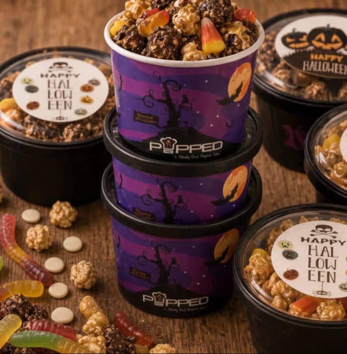 A styled product display of gourmet popcorn from popped las vegas on a rustic wooden table, surrounded by Halloween-themed ingredients like candy corn, gummy worms, pumpkin seeds, cinnamon, and mini pumpkins.