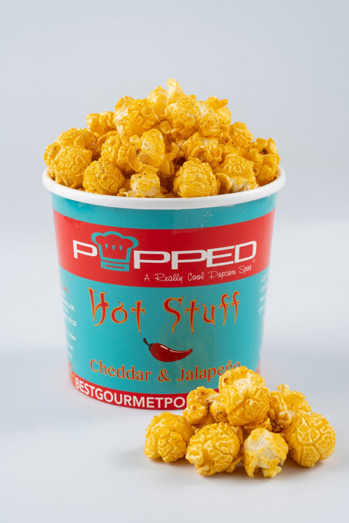 A cup of Popped Hot Stuff Cheddar & Jalapeño popcorn, with some popcorn spilled in front, on a light gray background.