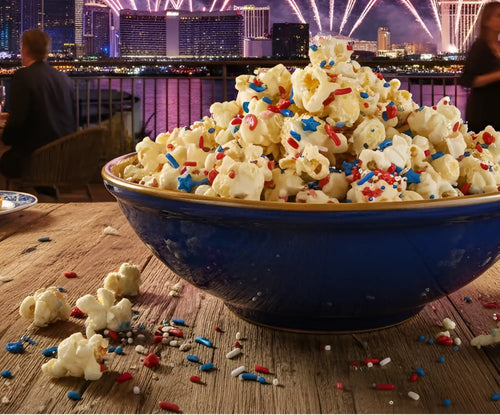 Large blue bowl of popcorn with colorful sprinkles on a wooden table, city skyline with fireworks in the background.