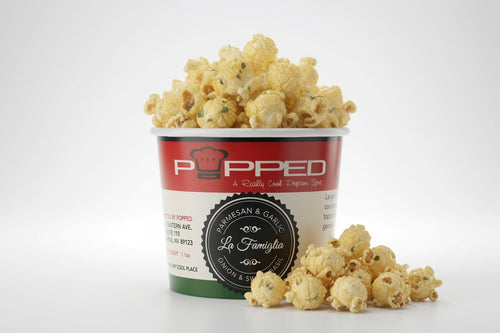 A cup of popcorn with some spilled around the base on a clean white background. The cup has a red, white, and green label with the brand name 'Popped' and a circular badge reading 'Parmesan & Garlic'.