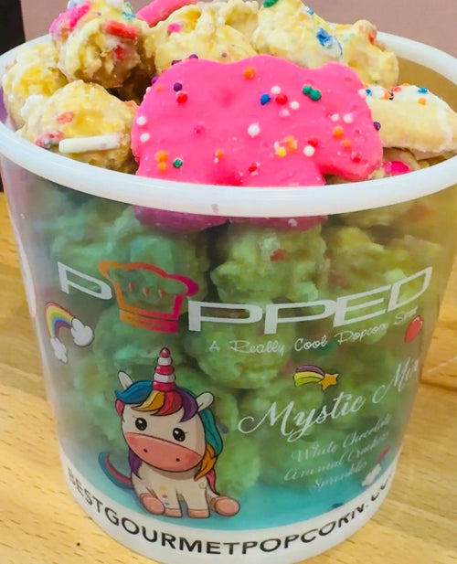 A clear plastic cup filled with colorful, sprinkle-covered popcorn and topped with a pink unicorn-shaped cookie.