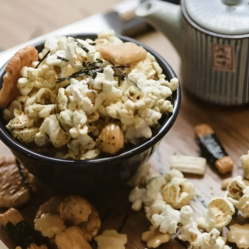 A black bowl overflowing with seasoned popcorn and various rice crackers, sprinkled with dried seaweed.
