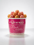 A cup of gourmet popcorn on a white background, featuring a pink paper cup labeled 'Popped Pink'adelic' with white chocolate raspberry popcorn visible at the top.