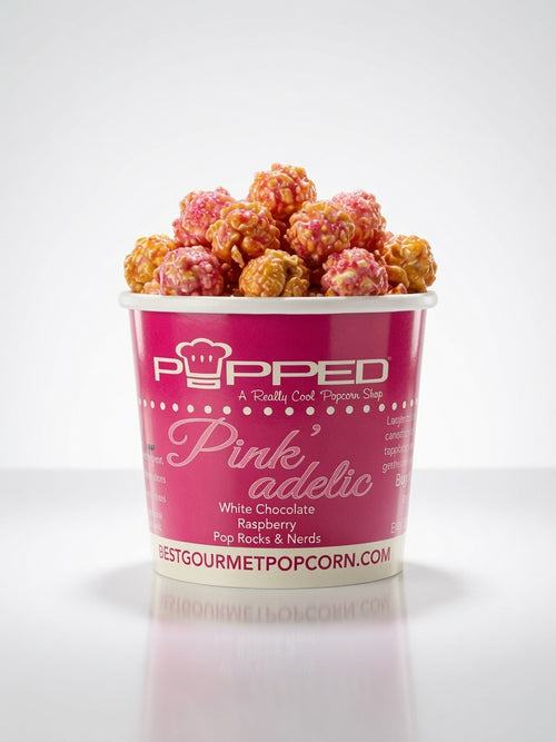 A cup of gourmet popcorn on a white background, featuring a pink paper cup labeled 'Popped Pink'adelic' with white chocolate raspberry popcorn visible at the top.