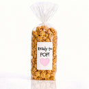 A clear cellophane bag filled with caramel popcorn and topped with a gathered twist, featuring a white label that reads 'Ready to POP!' with a pink heart on a clean white background. from popped las vegas