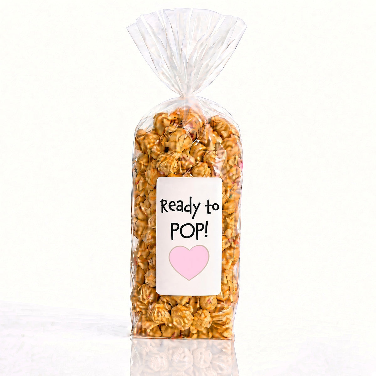 A clear cellophane bag filled with caramel popcorn and topped with a gathered twist, featuring a white label that reads 'Ready to POP!' with a pink heart on a clean white background. from popped las vegas