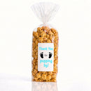 A clear cellophane bag filled with caramel popcorn, sealed and tied at the top, with a white printed label that reads 'Thank You for popping by!' in blue text with black baby footprints. The bag is photographed on a clean white background with a faint reflection below from popped las vegas