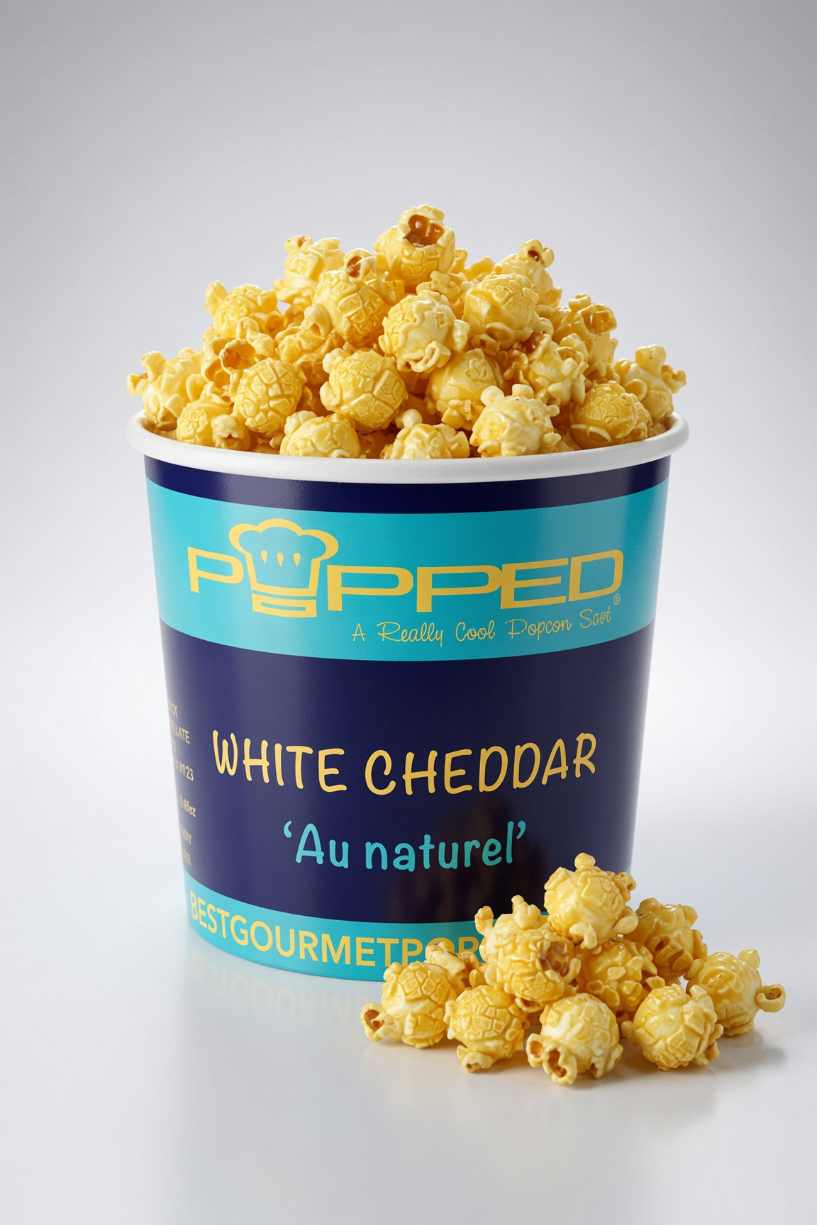 A bucket of Popped White Cheddar popcorn with a few pieces spilled in front, set against a clean light gray background.